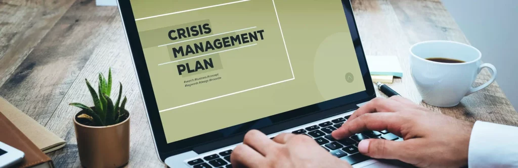 Cropped in photo of a man's hands typing on a laptop that reads "Crisis Management Plan" on the screen. Next to him is a plant and a mug of coffee.